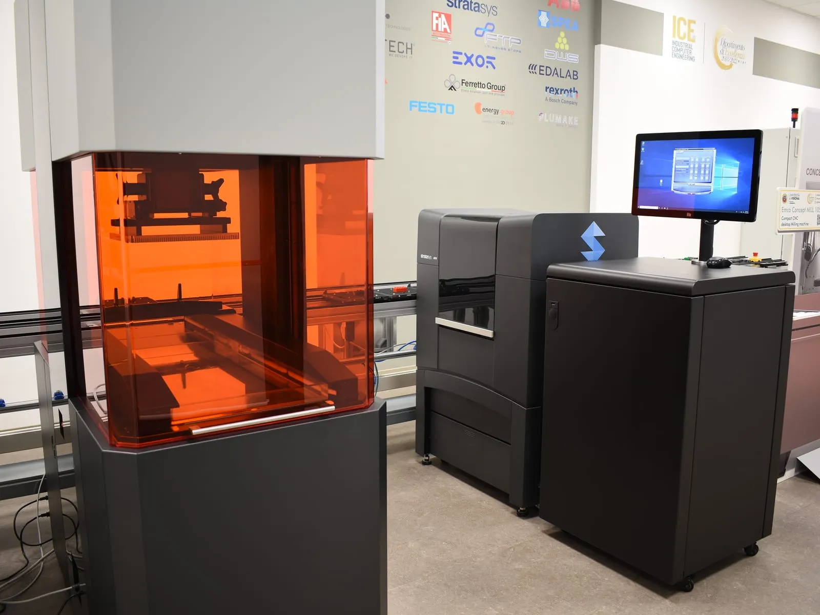 A resin 3D printer with an orange enclosure, next to an industrial printer and a control station