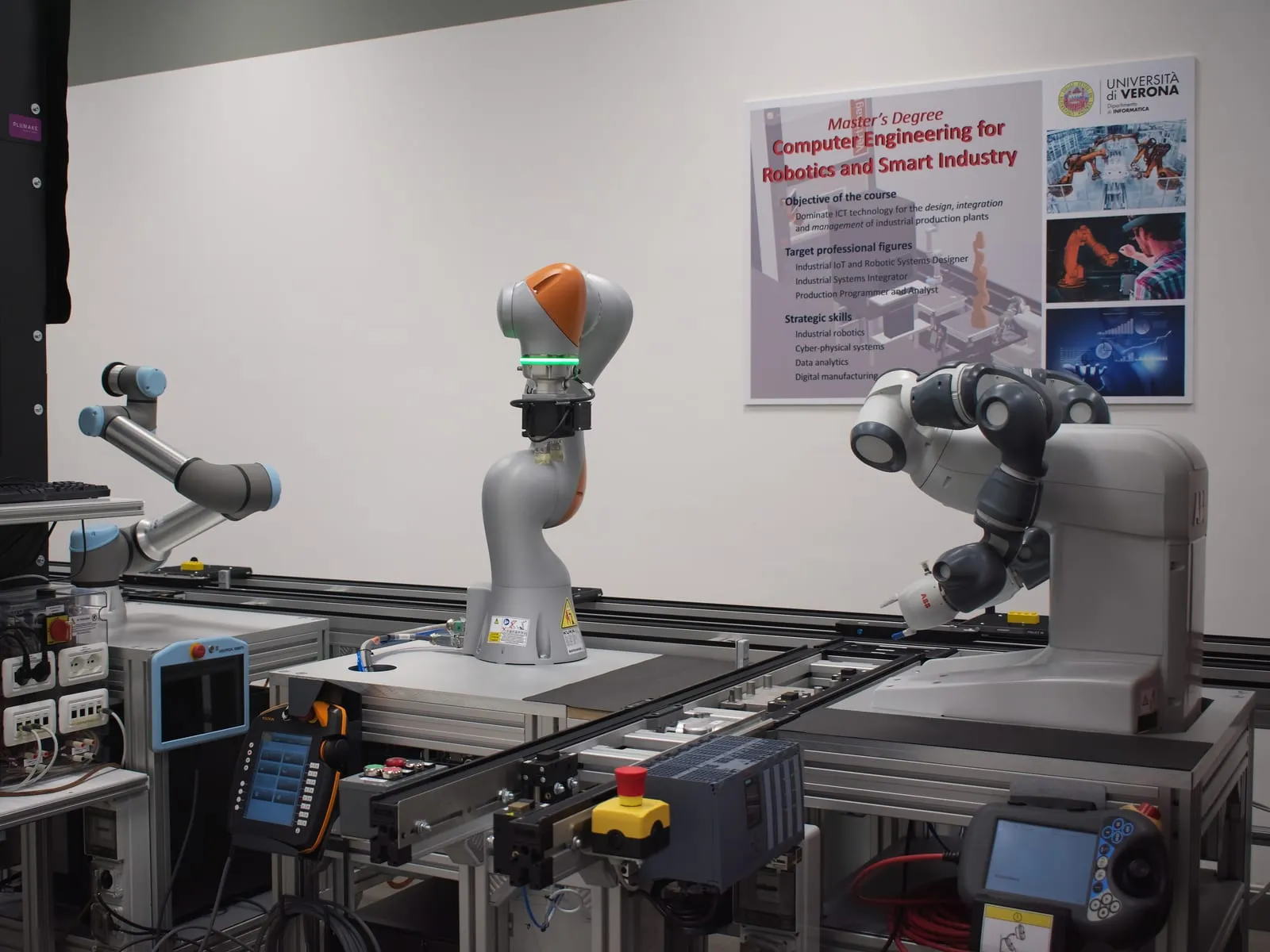 Two robot arms mounted over a conveyor, in front of a poster for the Computer Engineering for Robotics and Smart Industry degree